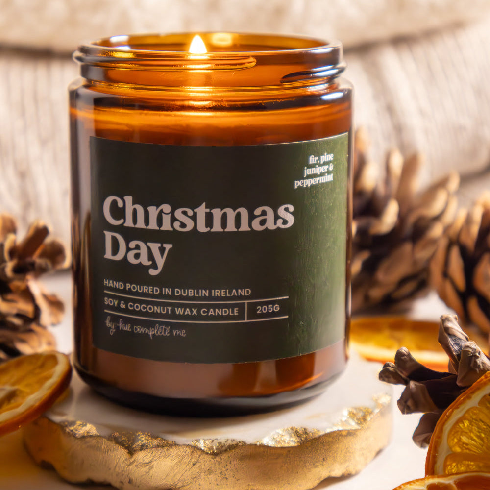 Candle in an amber jar labeled 'Christmas Day' surrounded by pine cones and dried oranges.