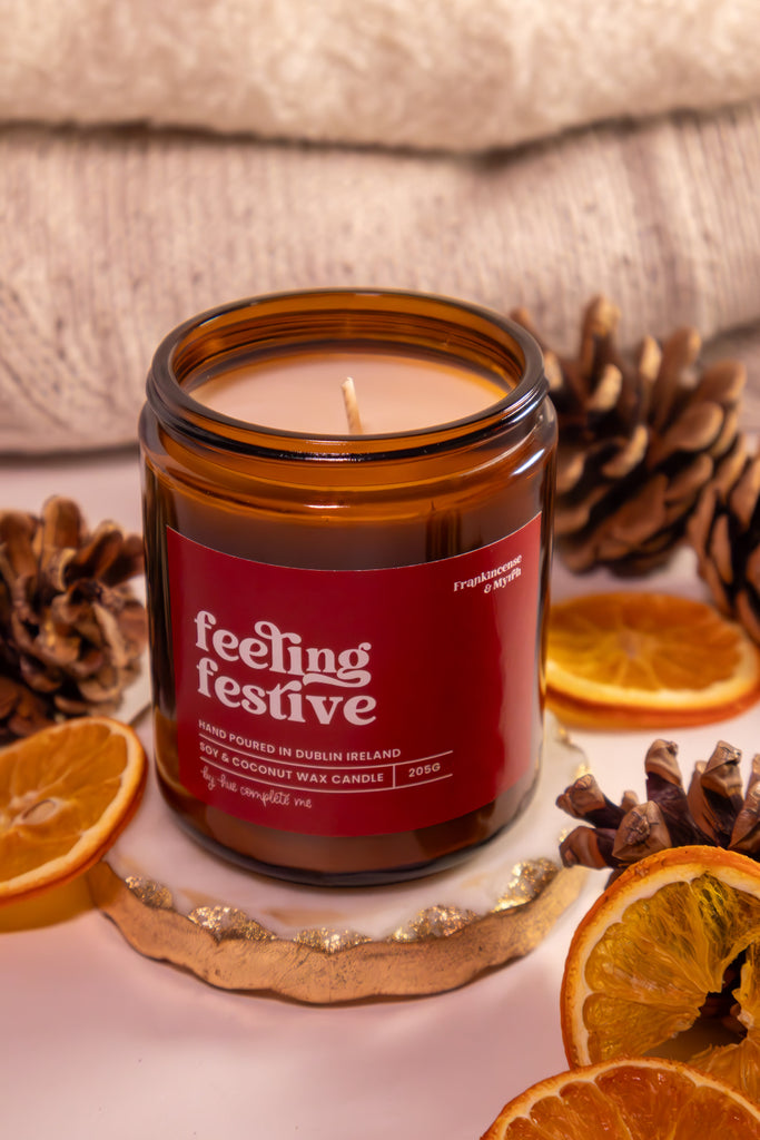Candle in an amber jar with a red label reading 'feeling festive' surrounded by pine cones and dried oranges.