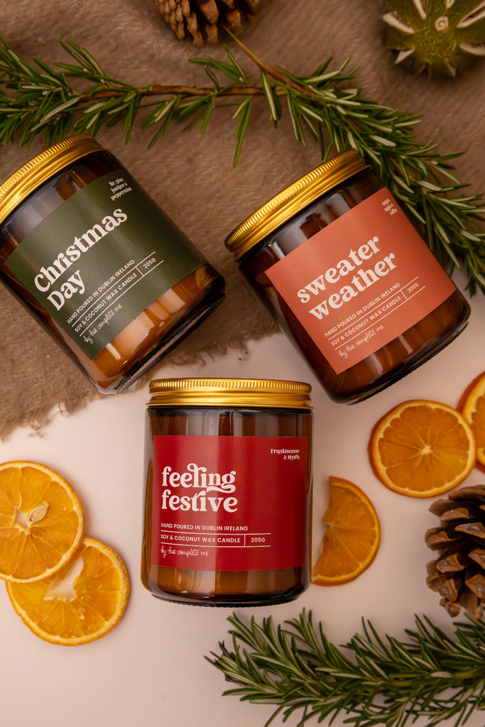 Three festive candles with labels 'Christmas Day', 'Sweater Weather', and 'Feeling Festive' surrounded by orange slices and pine branches.