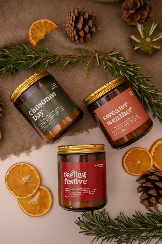 out three Christmas scented candles with festive labels surrounded by Christmas decorations on a wooden surface.