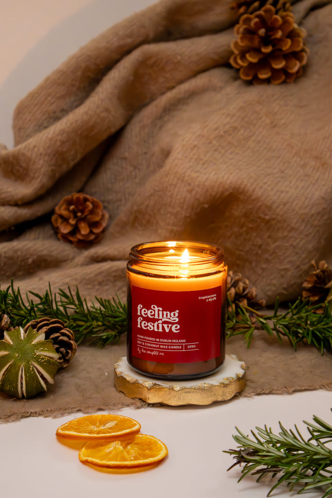 Candle labeled 'Feeling Festive' surrounded by pinecones, greenery, and a blanket on a neutral background