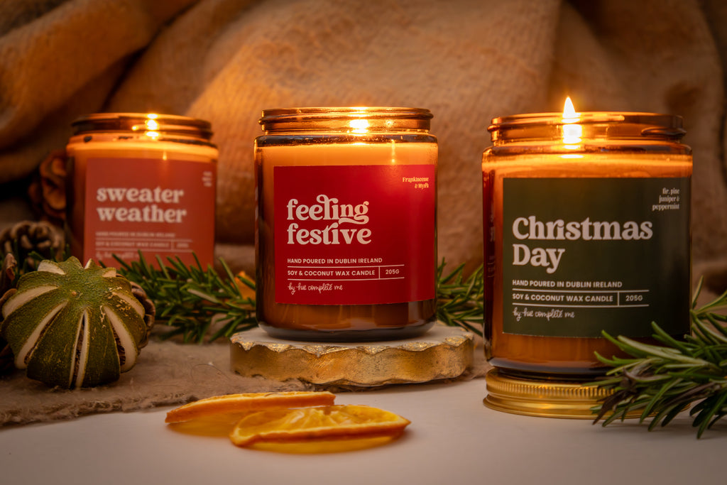 Three scented candles labeled 'sweater weather', 'feeling festive', and 'Christmas Day' on a decorative surface with greenery.